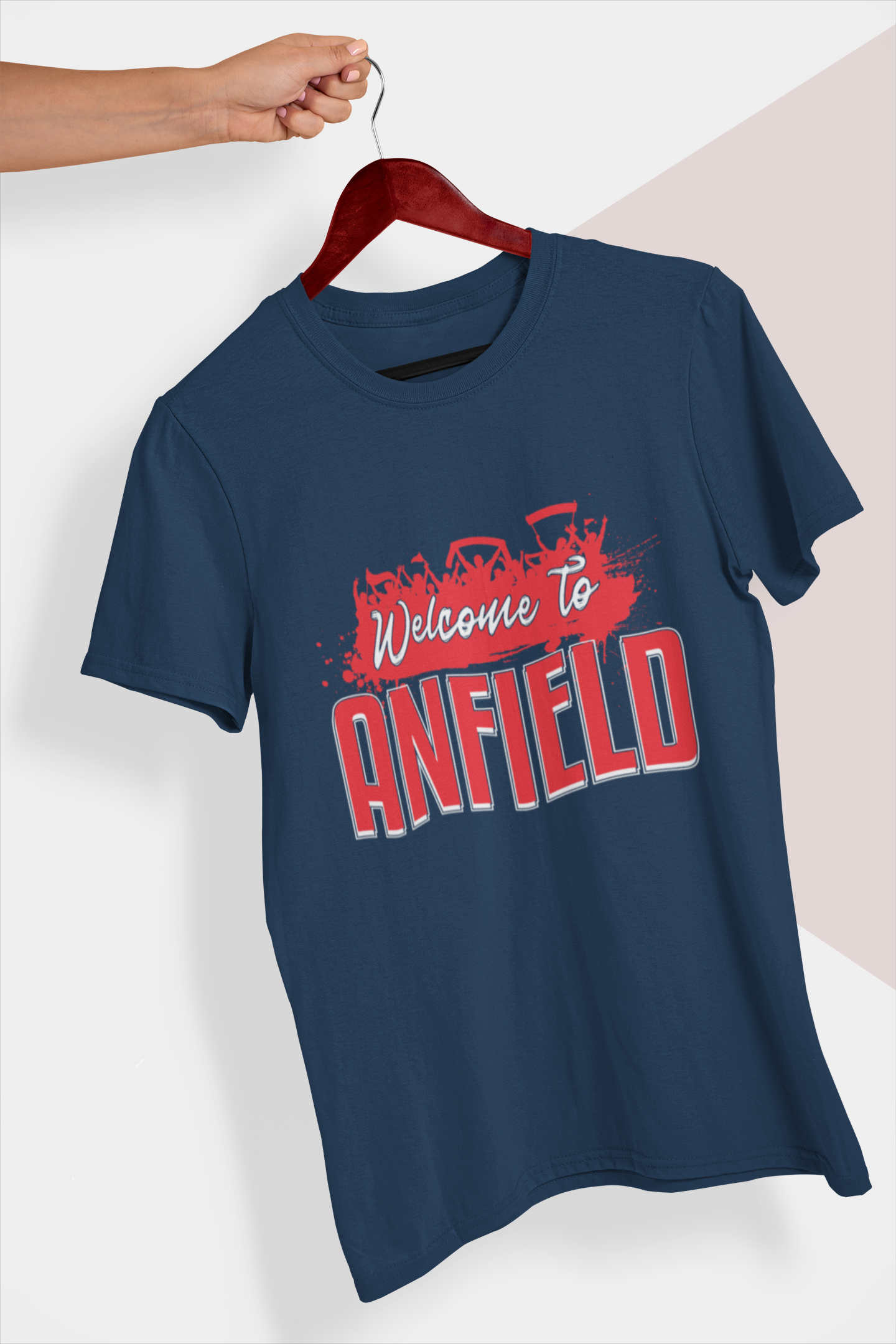Navy T-shirt on a hanger held in a person's hand in front of a white and cream colored background. Mock-up of a Welcome To Anfield Tee Shirt From Soccer Shocker With a Red and White Design