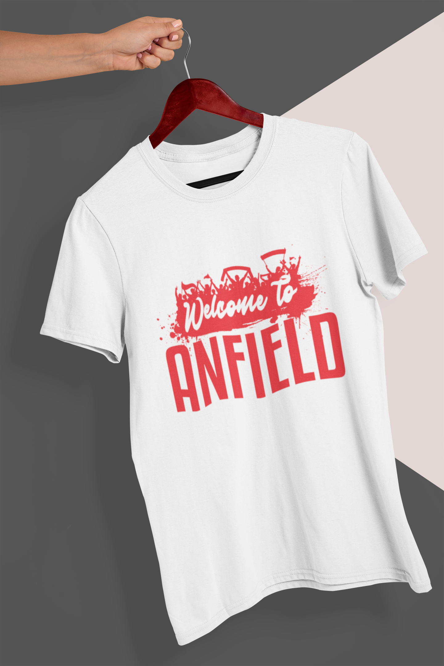 White tee shirt on a hanger held in a person's hand in front of a gray and cream colored background. Mock-up of a Welcome To Anfield Tee Shirt From Soccer Shocker With a Red and White Design